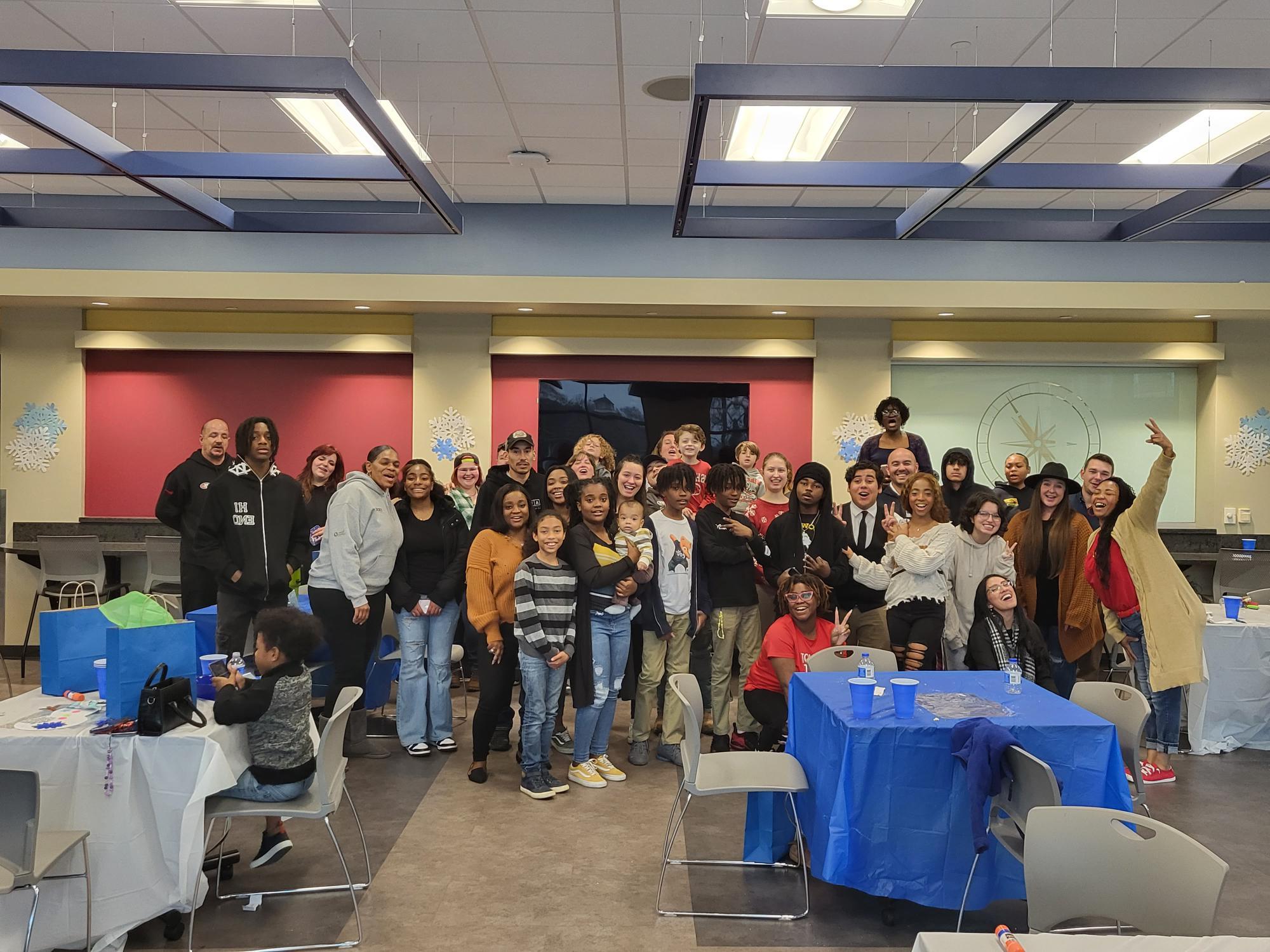 A diverse group of people posing for a group photo in a community room with tables covered in blue and white tablecloths. Decorative snowflakes are on the walls.